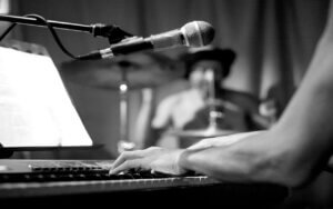 Photo en noir et blanc de mains sur un piano, avec un batteur en arrière fond.